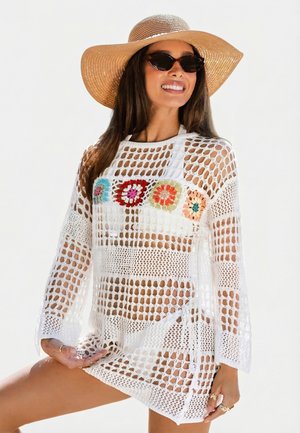 Frau mit breitkrempigem Hut, Sonnenbrille im Katzenaugenstil und weißem Häkelkleid mit bunten Blumenmustern, lächelnd und posierend.
