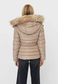 Beige gewatteerde jas met een capuchon afgezet met bont, een taille met riem en horizontale stiksels. Geweven stof, model van middelbare lengte en lange mouwen.