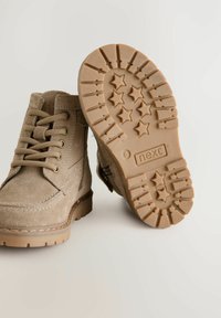 Beige suède enkellaarsjes met ronde neus, metalen vetertjes en veters. De buitenzool heeft een sterrenpatroon. Merkenlogo "next" zichtbaar op de zool.