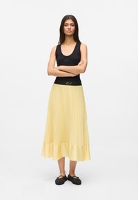 Jupe plissée jaune avec un ourlet en satin, associée à un débardeur noir avec un logo. Le modèle porte des chaussures noires en maille à enfiler.