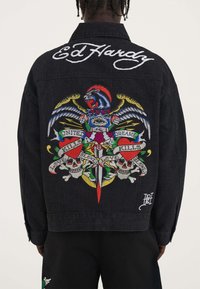 Giacca in denim nera con un grafico ricamato colorato che presenta un leone, un'aquila, spade, teschi e testo. Logo "Ed Hardy" in bianco in corsivo sul colletto.