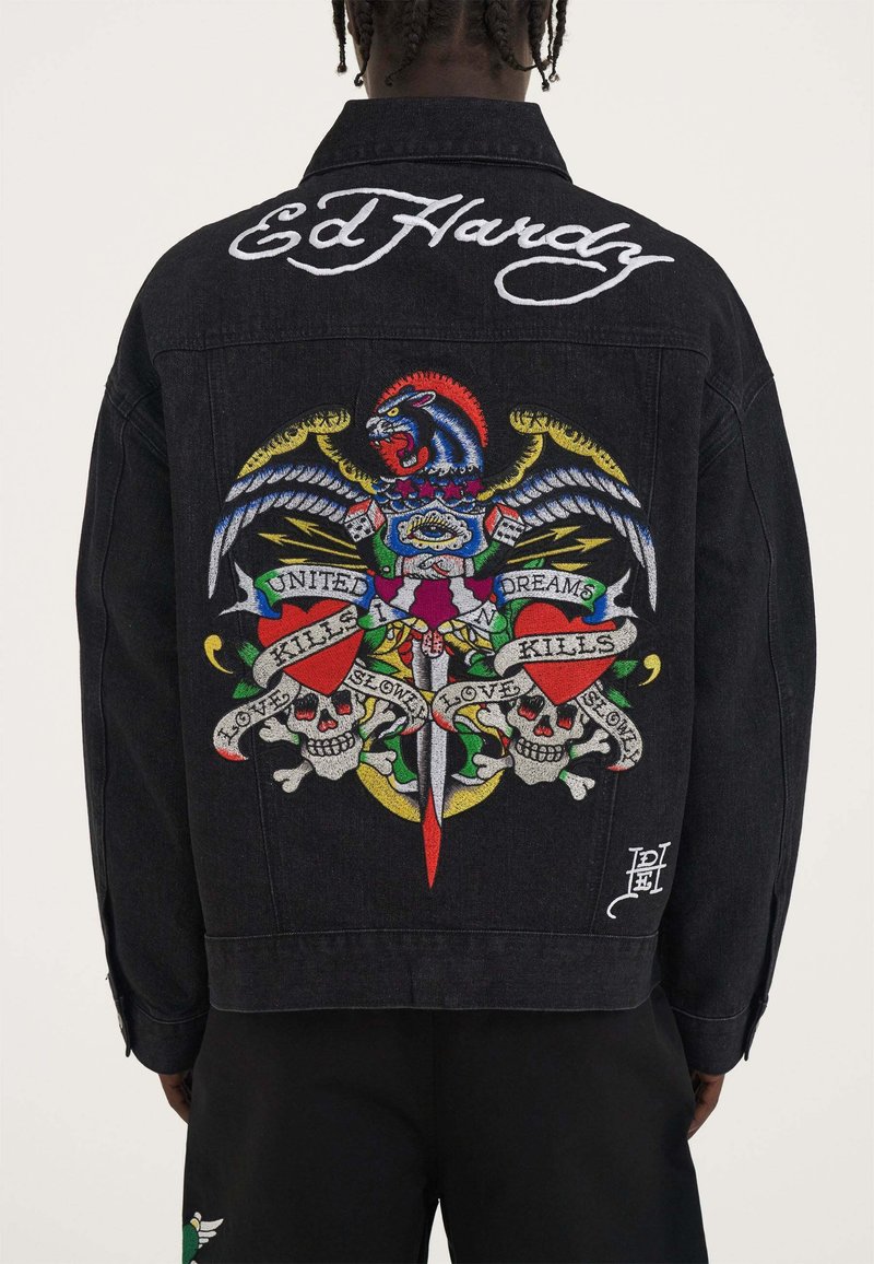 Giacca in denim nera con un grafico ricamato colorato che presenta un leone, un'aquila, spade, teschi e testo. Logo "Ed Hardy" in bianco in corsivo sul colletto.