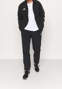 Černý adidas teplákový komplet s bundou na zip a bílými pruhy a logem, doplněný o odpovídající kalhoty, nošený s černými sportovními botami.