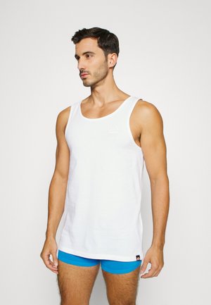 White tank top made of cotton, featuring a rounded neckline, slim fit, and a small logo on the chest. Worn with blue shorts.