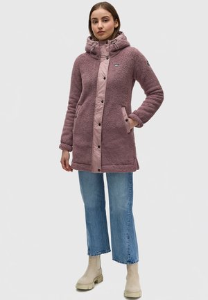 Manteau flou mauve avec un devant en nylon rose, boutons-pression et poches. Associé à un jean bleu clair légèrement court et des bottines beiges.