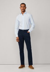 Camicia azzurra con bottoni e un motivo sottile abbinata a pantaloni blu scuro. Il modello sta in piedi con sicurezza con le mani in tasca, indossando scarpe marroni.