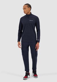 Giacca a zip blu navy con colletto alto, logo a contrasto sul petto, abbinata a pantaloni cargo blu navy e scarpe sportive nere-rosse.