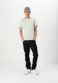Lichtgrijze t-shirt met een klein logo, donkerblauwe jeans en witte sneakers. Grijze pet maakt de outfit compleet. Effen achtergrond.