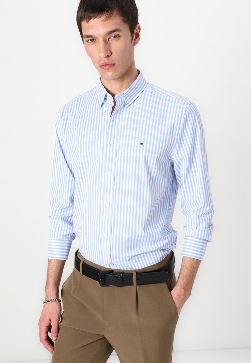 Chemise à rayures bleu clair et blanche, à boutons, avec un col, des manches longues et un petit logo sur la poitrine, portée avec un pantalon marron et une ceinture noire.