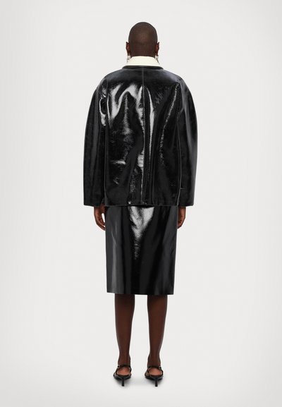 Giacca in pelle verniciata nera con colletto bianco, vestibilità oversize e cuciture visibili, abbinata a una gonna a tubino coordinata.