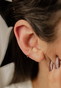 Goldene Creolen mit einem gedrehten Design, die eine Reihe kleiner klarer Edelsteine enthalten, die am Ohr einer Person getragen werden. Glatte Textur, einfaches Format.