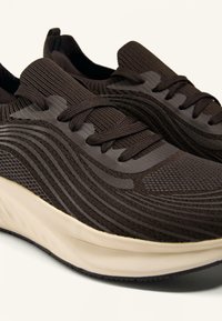 Donkerbruine sneakers met een textuur gebreid bovenwerk, platte veters en rubberen zool. Heeft golvende designelementen aan de zijkanten.