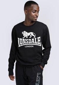 Fekete pulóver fehér oroszlán grafikával és "LONSDALE LONDON" felirattal. Kerek nyakú és hosszú ujjú. Puha anyagú textúra.