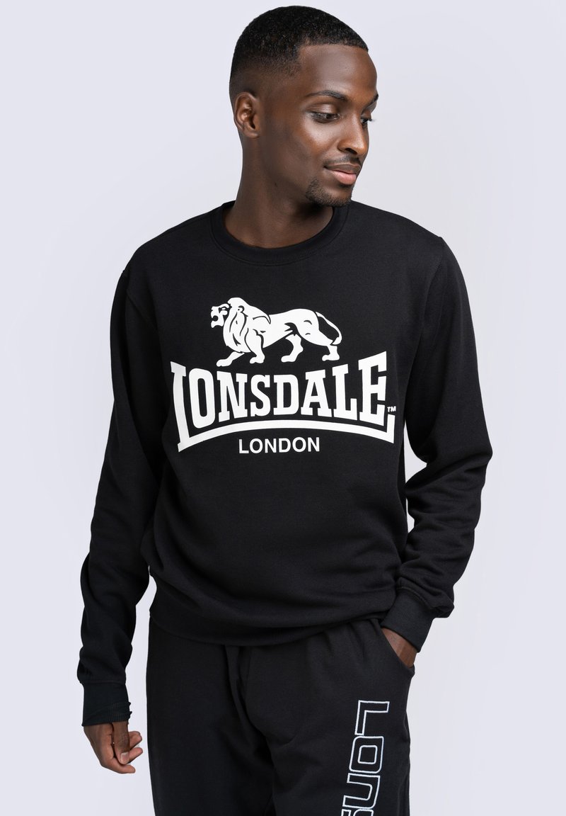 Fekete pulóver fehér oroszlán grafikával és "LONSDALE LONDON" felirattal. Kerek nyakú és hosszú ujjú. Puha anyagú textúra.