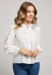 Chemisier blanc à boutons avec un col volanté agrémenté de broderies florales colorées. Manches longues bouffantes avec des poignets élastiques. Tissu doux.