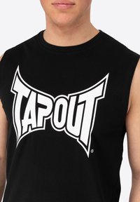 Fekete ujjatlan póló, ami pamutból készült, nagyméretű fehér "TAPOUT" logóval, merész, stilizált betűtípussal. Simább textúra, egyszerű dizájn.