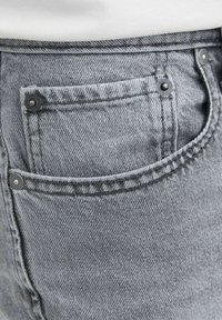 Szare dżinsy denimu z tylną kieszenią z kontrastowym przeszyciem i dwoma metalowymi nitami po bokach. Gładka tekstura z lekko wyblakłym wykończeniem.