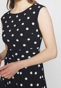 Ärmlös svart klänning med vita prickar, åtsittande midja, bärs av en person med ljus hy och brunt hår.