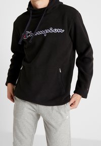 Felpa in pile nera con colletto alto, dotata di un logo ricamato in evidenza e tasche laterali con zip, abbinata a pantaloni della tuta grigi.