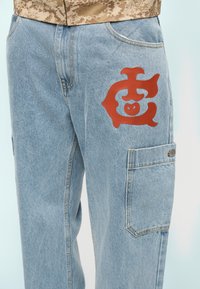 Φωτεινό γαλάζιο τζιν παντελόνι cargo με μεγάλη διπλωμένη τσέπη και κόκκινο αφηρημένο σύμβολο τυπωμένο στο δεξί μηρό.