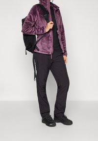 Person i lila fleecejacka, svarta byxor och vandringsskor som bär en svart ryggsäck, står mot en enkel ljus bakgrund.