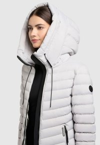 Lehká péřová bunda ve světle šedé barvě, s velkou, tvarovanou kapucí, horizontálním prošíváním, kapsami na zip a detaily se šňůrkami.