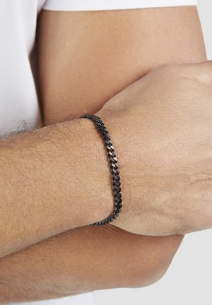 Männliche verschränkte Arme mit einem schwarzen Metallgliederarmband am Handgelenk vor einem weißen Hemdhintergrund.