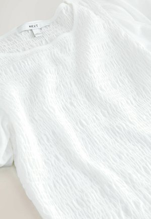 Balta virskrekls ar teksturētu virsmu un viļņotu rakstu. Audums šķiet viegls, ar īsām piedurknēm un apaļu kakla izgriezumu.