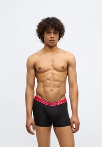 Young man with curly hair wearing black boxer briefs with red waistband labeled "Tommy Hilfiger" standing against plain background.