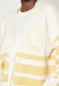 Cardigan bianco con strisce gialle, scollatura a V e chiusura a bottoni. Presenta un logo ricamato a forma di "G" e dettagli testurizzati sul tessuto.