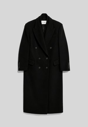 Femme aux cheveux bruns mi-longs portant un long manteau noir croisé sur un pantalon noir à jambes larges et un haut blanc, debout devant un fond uni.