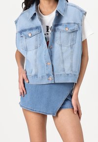 Light blue denim vest with oversized fit, featuring two chest pockets, silver buttons, and a slightly frayed hem. Paired with a blue mini skirt.