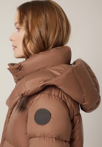 Brązowa kurtka puchowa z wysokim kołnierzem, z okrągłą naszywką z logo na lewym ramieniu oraz błyszczącą, pikowaną fakturą.