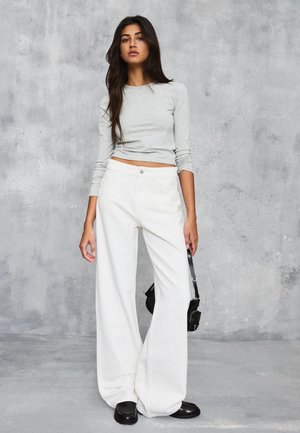 Ženska z dolgimi temnimi lasmi, oblečena v svetlo sivo dolgoročno oblečeno majico in široke bele hlače, drži majhno črno torbico.