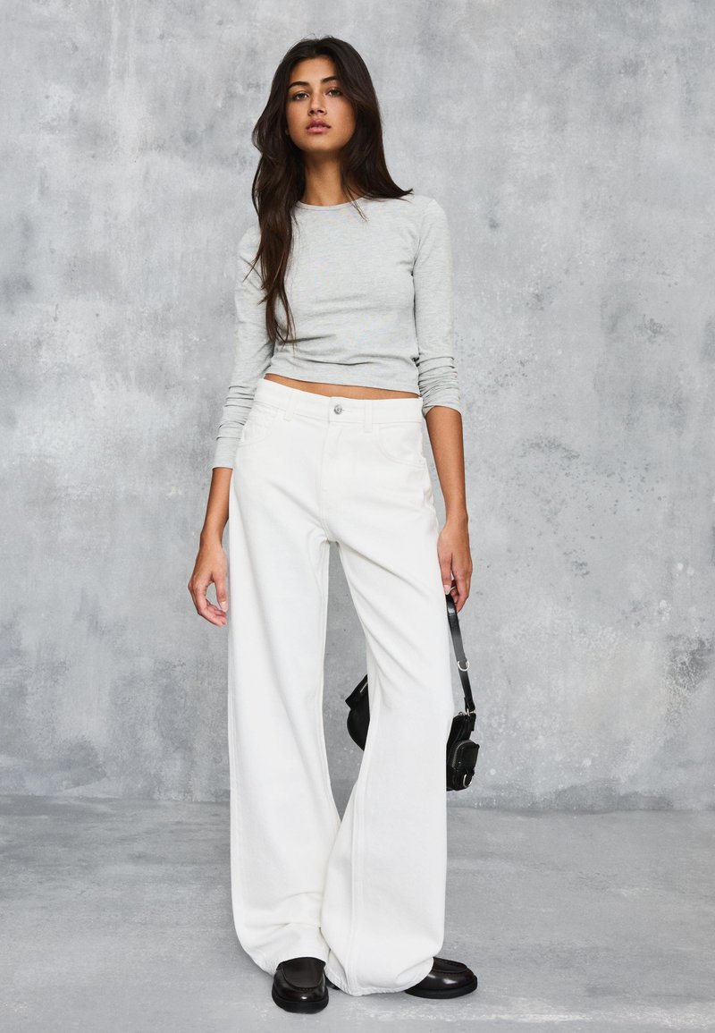 Femme aux longs cheveux foncés portant un crop top à manches longues gris clair et un pantalon blanc à jambes larges, tenant un petit sac à main noir.