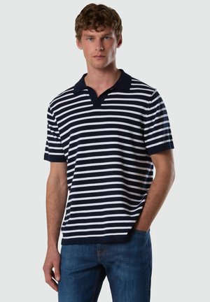 Giovane uomo con capelli ricci che indossa una polo a maniche corte a righe orizzontali blu navy e bianco e jeans blu, in piedi con una mano in tasca.