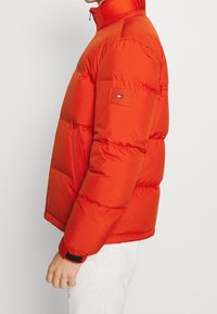 Sido vy av en person som bär en ljus röd vadderad jacka med dragkedja och en liten logotyp-patch på överarmen.