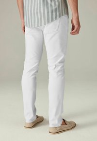 Weiße Baumwollhosen mit schmaler Passform, von hinten gezeigt. Dazu leichte, hellbraune Slipper mit geflochtenen Details.