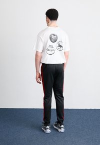 Vit t-shirt med korta ärmar och tryck på baksidan, kombinerad med svarta byxor med röda sidostripe; svarta sportskor.