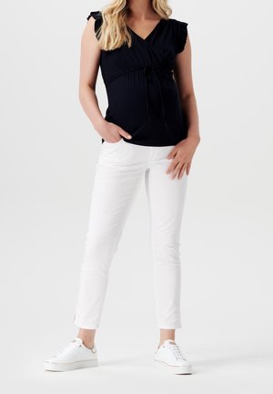 Femme portant un débardeur noir et un pantalon blanc avec des baskets blanches, debout avec une main dans la poche sur un fond uni.