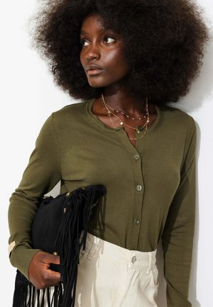 Femme aux grands cheveux bouclés portant un pull boutonné vert olive, des colliers dorés superposés, un pantalon blanc et tenant une pochette noire à franges.