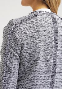 Kvinna klädd i en gråvit tweedjacka med struktur och svartvitt fransdetaljering på ryggen och axelsömmarna.