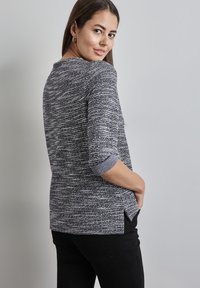 Grauer und schwarzer strukturierter Pullover mit Dreiviertelärmeln, umgeschlagenen Bündchen und Seitenschlitzen, kombiniert mit schwarzen Hosen.