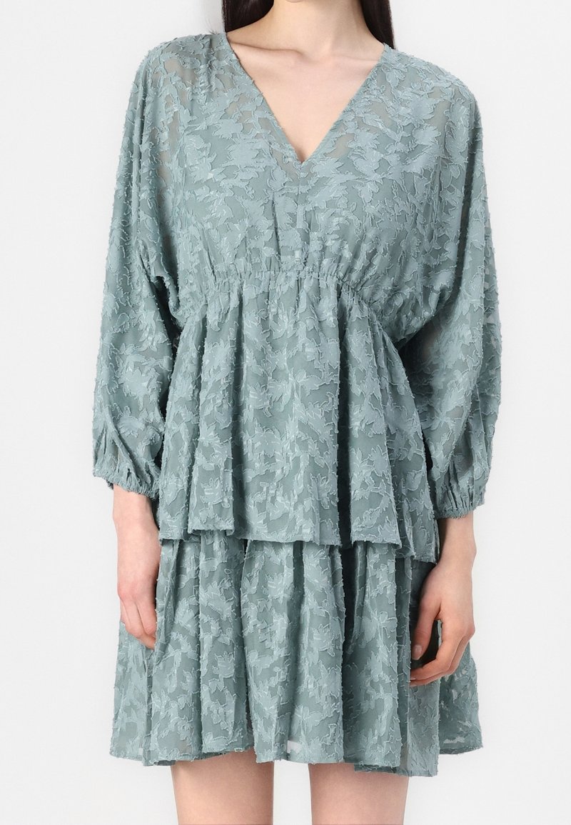 Femme portant une robe en dentelle bleu clair avec manches longues, col en V, taille cintrée et jupe superposée longueur genoux.