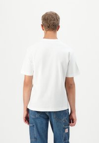 Person med kort, krøllet hår iført en simpel hvid T-shirt med korte ærmer og blå denimbukser, som vender væk på hvid baggrund.