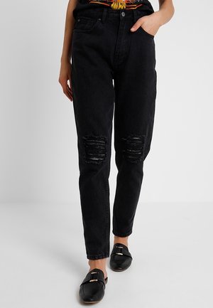 Kvinde iført sorte jeans med høj talje og slidte knæ, en indstukket T-shirt med grafik og sorte slip-on loafers med guldlåse.