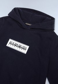 Námornícky modrá mikina s kapucňou vyrobená z jemného materiálu, s veľkým bielym obdĺžnikovým logom a čiernym textom. Obsahuje priestrannú kapucňu a voľný strih.