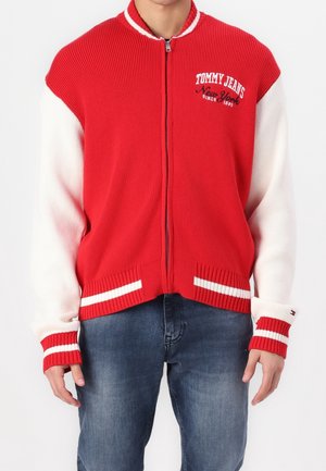 Mann trägt einen roten Zip-up-Pullover mit weißen Ärmeln und dem Logo „Tommy Jeans New York“ auf der Brust, kombiniert mit blauen Jeans.