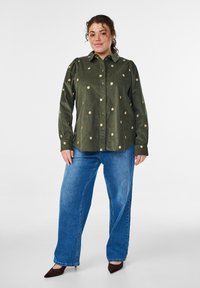 Grünes Hemd mit Knöpfen und Blumenstickerei, kombiniert mit blauen Jeans mit weitem Bein und braunen spitzen High Heels. Das Hemd hat einen Kragen und geraffte Ärmel.