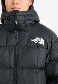 Čierna puffová bunda s vysokým golierom, má predný zips a prešívané detaily. Logo The North Face je výrazne zobrazené v bielej farbe.